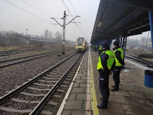 Policjanci w odblaskowych kamizelkach stoją na peronie. W tle widać nadjeżdżający pociąg. Pogoda zimowa. Pochmurne niebo.