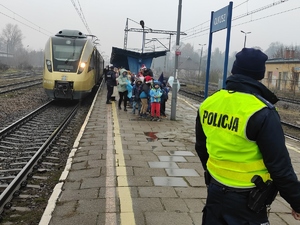 Z pociągu wysiadają dzieci i opiekunowie. Po prawej stronie widać policjanta w odblaskowej kamizele. Policjant stoi tyłem do zdjęcia, na plecach napis POLICJA. Pogoda deszczowa pochmurne niebo.