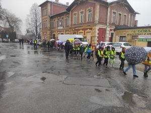 Duża grupa dzieci idzie w parach jeden zza drugim. Niektóre dzieci mają ubrane kamizelki odblaskowe, w rękach trzymają parasolki. W tle budynek PKP w Olkuszu. Pogoda deszczowa pochmurne niebo.