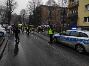 Policjanci stoją na ulicy blokując przejazd samochodom. Duża grupa dzieci przechodzi na przejściu dla pieszych, jeden za drugim. Po prawej radiowóz na sygnałach. W tle policjant ruchu drogowego w białej czapce i samochód na sygnałach. Pogoda deszczowa. Jezdnia mokra. Po obu stronach jezdni chodniki i zabudowania.