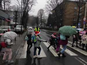 Grupa dzieci pod parasolkami przechodni przez przejście dla pieszych. Na jezdni stoi policjant ruchu drogowego, który  kieruje ruchem. W tle na jezdni  samochody. Po obu stronach jezdni chodniki i zabudowania.