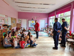 Ratownik rozmawia z dziećmi. Dzieci siedzą na dywanie w kole. Po prawej stronie stoją dwaj policjanci. Zajęcia odbywają się w klasie.