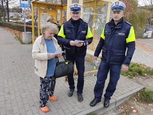 Dwaj policjanci ruchu drogowego (białe czapki) w odblaskowych kamizelka przekazują seniorce odblask i ulotki profilaktyczne. Zdjęcie zrobione w pobliżu wiaty przystankowej. W tle budynek, parking i samochody.