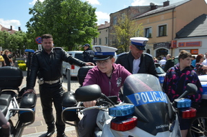 Chłopiec na motocyklu policyjnym w białej czapce policjantka ruchu drogowego. W tle dwóch policjantów. Zdjęcie wykonane na olkuskim Rynku. Pogoda słoneczna.