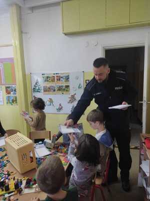 Dzielnicowy w przedszkolu rozdaje przedszkolakom kartki edukacyjne .Dzieci stoją przy policjancie. Zdjęcie zrobione  klasie , w tle zabawki i klocki.