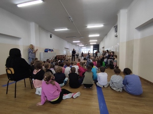 Policjantka prowadzi pogadankę z uczniami Szkoły Podstawowej nr 2 w Olkusz. Spotkanie w korytarzu szkolnym. Duża grupa dzieci klas 1-3 siedzi na podłodze. Po środku stoi policjantka.