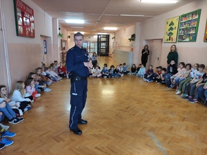Policjant – dzielnicowy prowadzi spotkanie z dziećmi na szkolnym korytarzu. Dzieci siedzą w kręgu. Po środku stoi policjant.