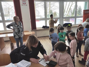 Policjantka wykonuje odciski palców przedszkolakom. Grupa dzieci stoi przy policjantce. W tle widać pozostałe dzieci i funkcjonariusza Straży  Pożarnej.