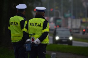 Dwóch policjantów ruchu drogowego (białe czapki) w odblaskowych kamizelkach prowadzi nadzór na drodze. W tle widać ruch uliczny.