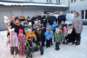 Grupa przedszkolaków wraz z opiekunami oraz policjantem i dwoma funkcjonariuszami straży miejskiej na tle budynku komisariatu i pojazdów służbowych straży i policji pozują do zdjęcia. Pogoda zimowa (śnieg).