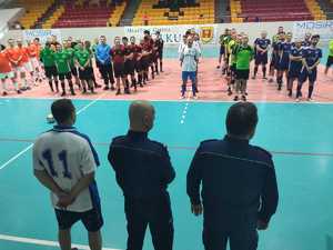 Komendant olkuskiej Policji  i Naczelnik sztabu wraz z uczestnikiem turnieju  stoją przodem do drużyn. Drużyny ustawione w rzędach. Zdjęcie w hali sportowej. W tle trybuny.