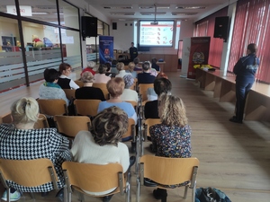 Strażak podczas prelekcji z seniorami. W tle slajd prezentacji. Po prawej stronie stoi policjantka przodem do strażaka. Na zdjęciu widać osoby siedzące w rzędach na stołkach.