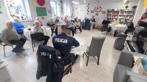 W sali środowiskowego Domu Pomocy Społecznej, policjant siedzi przodem do seniorów i prowadzi prelekcje. Seniorzy siedzą przy stoliczkach.