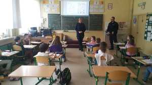 Policjantka i policjant na spotkaniu w klasie z dziećmi. Dzieci siedzą w szkolnych ławkach na wprost policjantów.