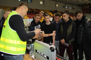 Technik kryminalistyki w odblaskowej kamizelce na słoiku ujawnia ślady linii papilarnych. Grupa gromadzi się przed policjantem.