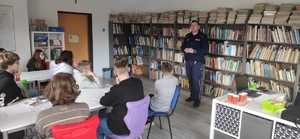 Dzielnicowy z Komendy Powiatowej Policji w Olkuszu prowadzi zajęcia z uczniami klasy VIII ze Szkoły Podstawowej w Sienicznie. Zajęcia w klasie bibliotecznej. Dzieci siedzą w szkolnych ławkach. W tle półki z książkami.