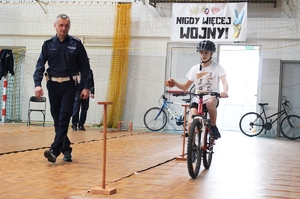 Uczestnik turnieju na rowerze przy pierwszej przeszkodzie na torze oraz policjant obserwujący sposób wykonania zadania.