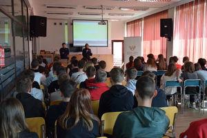 Policjantka i policjant na prelekcji z uczniami klas 8. Młodzież siedzi na stołkach w rzędach. W tle widać prezentacje multimedialna.