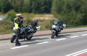 Policjant ruchu drogowego (biała czapka) w odblaskowej kamizelce na poboczu drogi dokonuje pomiaru prędkości kierujących. Obok dwa policyjne motocykle.