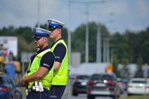 Dwóch policjantów ruchu drogowego (białe czapki, kamizelki odblaskowe) prowadzi nadzór na drodze.