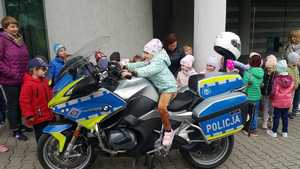 Dziewczynka siedzi na policyjnym motocyklu, wokół niej grupa przedszkolaków oraz trzy opiekunki dzieci.