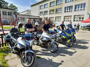 4 policjantów Wydziału  Ruchu Drogowego przy policyjnych  motocyklach. W tle budynek Szkoły Podstawowej nr 4 w Olkuszu. Pogoda letnia, bezchmurne niebo.