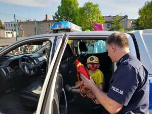 Policjant soi przy radiowozie i pokazuje dziecku rzutkę. Dziecko siedzi na tylnym siedzeniu . Ma pomalowaną na czerwono twarz.