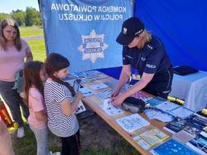 Policjantka na stanowisku policyjnym wykonuje dzieciom odbitki palców. Na stoliku ulotki, materiały edukacyjne, książeczki, broszury, malowanki.