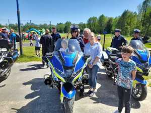 dziecko siedzi na motocyklu obok policjantka ruchu drogowego oraz organizator imprezy. W tle krajobraz oraz ludzie. Pogoda słoneczna