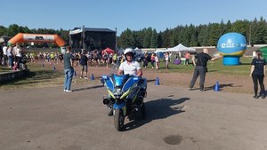 Policjant ruchu drogowego na motocyklu podczas zabezpieczenia biegu ulicznego. W tle meta i stadion sportowy oraz  uczestnicy imprezy.