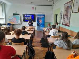 Policjant na spotkaniu w klasie  z uczniami Zespołu Szkół w Wolbromiu. Uczniowie siedzą w szkolnych ławkach. Przed nimi stoi policjant. W tle baner Komendy Powiatowej Policji w Olkuszu