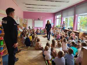 Policjantka wraz z policjantem w klasie szkolnej  na spotkaniu z dziećmi. Policjantka stoi wśród dużej grupy dzieci. Dzieci siedzą na podłodze tworząc krąg dookoła policjantki