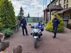 Dwaj policjanci ruchu drogowego stoją pomiędzy policyjnym motocyklem. Na motorze siedzi przedszkolak. Przed policjantami stoi grupa przedszkolaków. Obok po prawej stronie na zdjęciu, widać część budynku szkolnego.