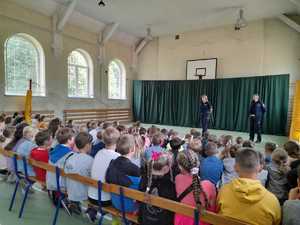 Policjantka i policjant na spotkaniu z uczniami szkoły Podstawowej nr 1 w Olkuszu. Spotkanie na sali gimnastycznej. Policjanci stoją  przed liczna grupą uczniów, którzy siedzą na ławeczkach.