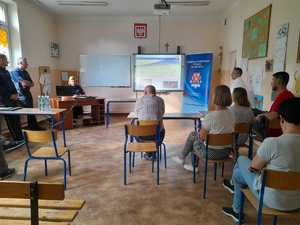 Debata społeczna w Sienicznie prowadzona w szkolnej sali lekcyjnej. Uczestnicy siedzą na stołkach przodem do policjantki. na przeciwko policjantka  prowadzi debatę. Po jej prawie stronie slajd na projektorze. Obok  rollup i logo Komendy Powiatowej Policji w Olkuszu wraz z danymi teleadresowymi.