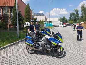 Dwaj policjanci ruchu drogowego przy motocyklach. Na terenie obiektu sportowego, gdzie odbywają się zawody sumo. W tle obiekt sportowy, ulica i zaparkowane auta.