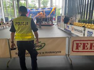 Policjant w odblaskowej kamizelce podczas zabezpieczenia imprezy sportowej. W tle, w hali sportowej,  na podium zawodnicy