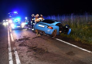 Samochód po wypadku. Auto znajduje się częściowo w rowie. Tylne koła na jezdni. W tle pojazdy straży pożarnej i policji na błyskach. Na drodze przy pojeździe stoją  strażacy. Zdjęcie nocą.