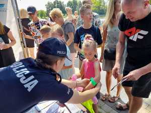 Policjantka pochyla się nad dziewczynka i zakłada jej na rękę odblask. Na stole kolorowe broszury gadżety i ulotki. W tle gromadzą się ludzie i dzieci.