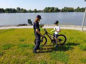 Policjantka podaje rowerzyście kamizelkę odblaskowa w tle zalew i pejzaż.