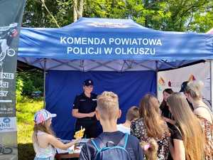 Policjantka przy stanowisku policyjnym przy namiocie rozdaje dzieciom gadżety. Wokół stanowiska gromadzą się rodziny.