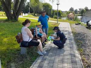 Policjantka pochyla się na dziećmi i wręcza książeczki. Dzieci siedzą z rodzicami na ławce. W tle zalew i pejzaż.