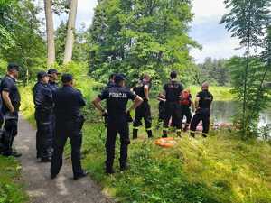 Policjanci i strażacy nad Stawem Kwaśniowskim. Na stawie ponton strażacki wraz z e strażakiem. Wodowanie pontonu. Dokoła pejzaż pogoda letnia.