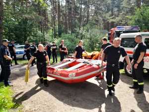 Strażacy niosą ponton strażacki. W tle policjanci i wozy strażackie i radiowozy. W oddali pejzaż.