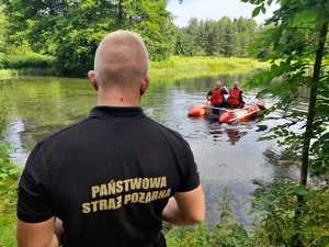 Strażak stoi przed stawem. na stawie ponton strażacki z dwoma strażakami w kamizelkach ratowniczych. Dokoła zieleń i drzewa.