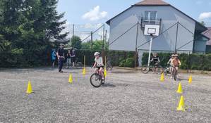 Dziewczynka na rowerze w kasku ochronnym pokonuje tor przeszkód. Dziewczynka jedzie pomiędzy pachołkami. W tle stoi policjant ruchu drogowego (biała czapka), dzieci na rowerach przygotowują się do jazdy. W oddali budynek.