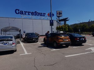 Policjant na parkingu przy centrum handlowym zagląda do samochodu przez okno. Policjant sprawdza  czy w zaparkowanym pojeździe nie znajdują się pozostawione osoby lub zwierzęta. Pogoda letnia bezchmurne niebo. W tle budynek, parking i zaparkowane samochody