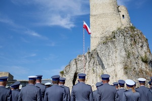 Policjanci w galowych mundurach stoją tyłem. Przed nimi powiewa flaga na maszcie. W oddali wieża zamku. Pogoda letnia, bezchmurne niebo.