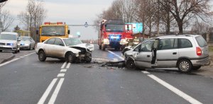 Dwa samochody osobowe marki opel zafira i audi  po zderzeniu czołowym. Przody obu pojazdów zniszczone. na drodze znajdują się elementy zderzaków, kołpaki i inne części. Droga zablokowana przez te dwa pojazdy. W tle widać dwa nieoznakowane samochody policyjne z włączonymi sygnałami świetlnymi oraz samochód strażacki.