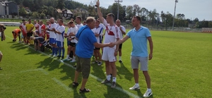 Prezes Ludowego Klubu Sportowego Kłos gratuluje piłkarzowi. W tle boisko i zawodnicy stojący w rzędzie